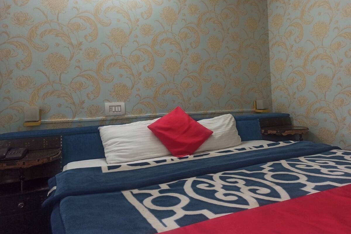 Quarto de hotel com papel de parede floral, cama com colcha de padrão azul e branco, travesseiro vermelho e mesa de cabeceira com telefone.