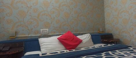 Una habitación de hotel con papel tapiz floral, una cama con colcha de rayas azules y blancas, un almohadón rojo y una mesita de noche con un teléfono.