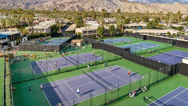 A tennis court complex with multiple courts, surrounded by residential buildings and palm trees, with mountains in the background.