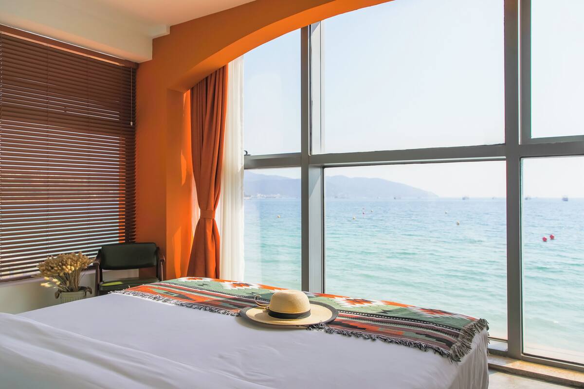 Een hotelkamer met een groot raam dat uitzicht biedt op de zee en verre bergen.