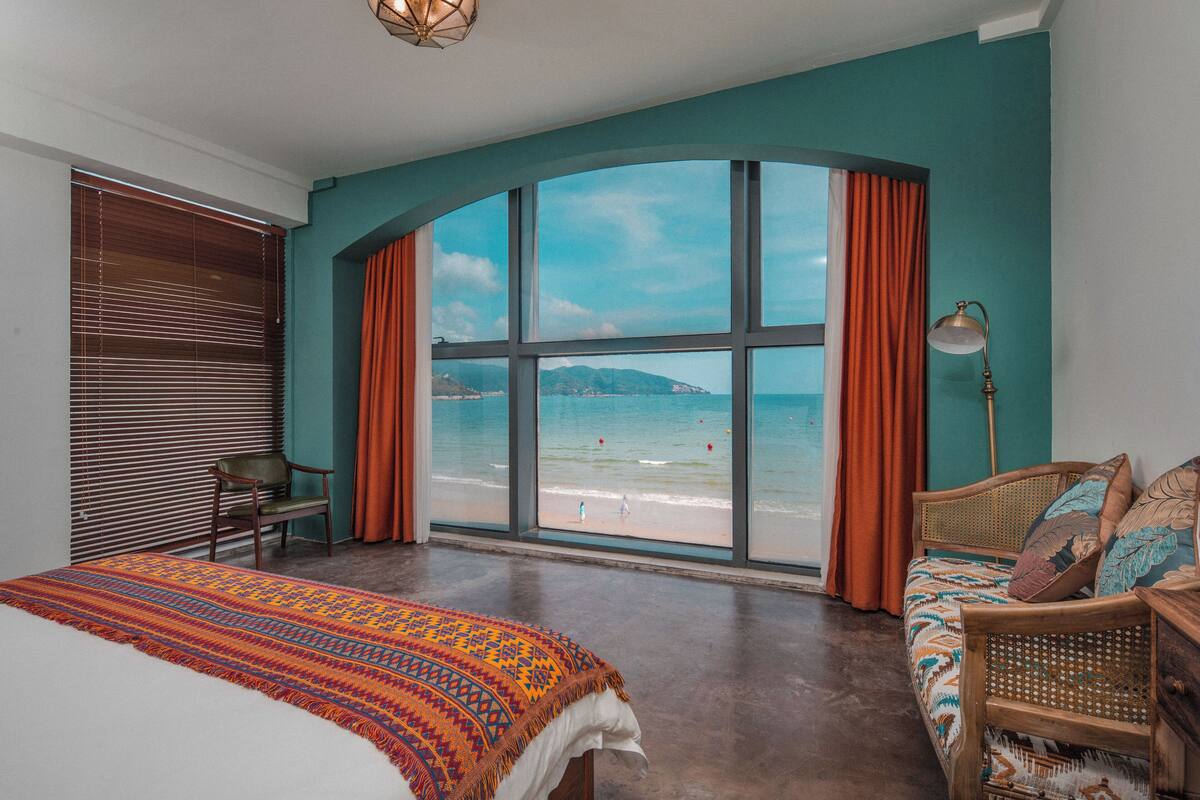 Een slaapkamer met een groot raam met uitzicht op een strand, een bed, een stoel en een nachtkastje.