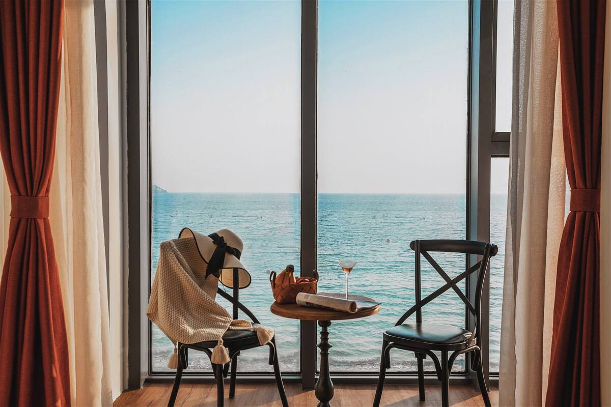 Een kamer met een groot raam met uitzicht op zee, twee stoelen, een kleine tafel met een drankje en een tas, en een zicht op de kustlijn.