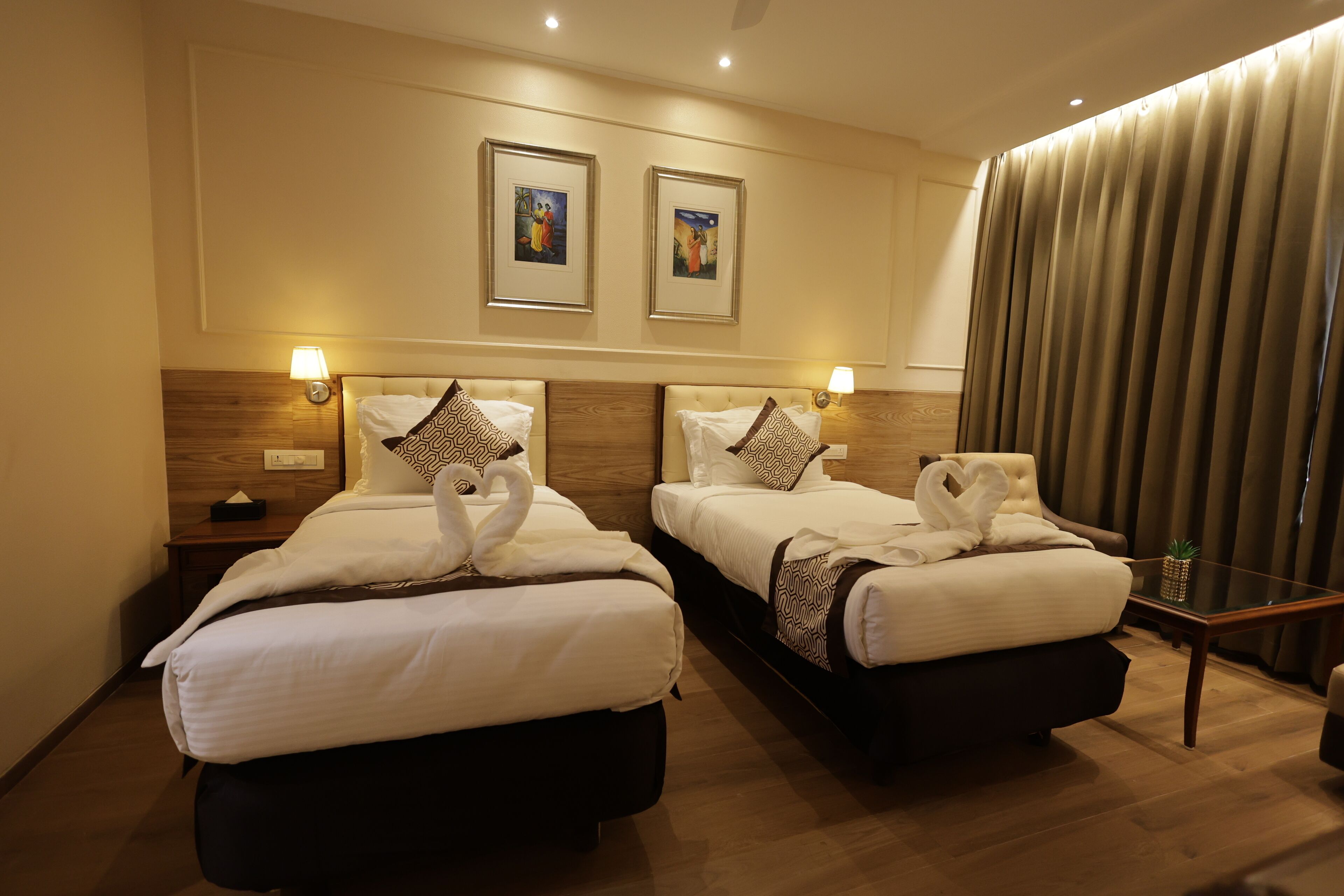 A hotel room with two beds, each with a towel swan decoration, framed artwork on the wall, and a small table with a plant.