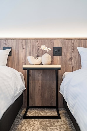 Quarto com duas camas, uma mesinha de cabeceira com um vaso e um quadro de interruptores na parede.