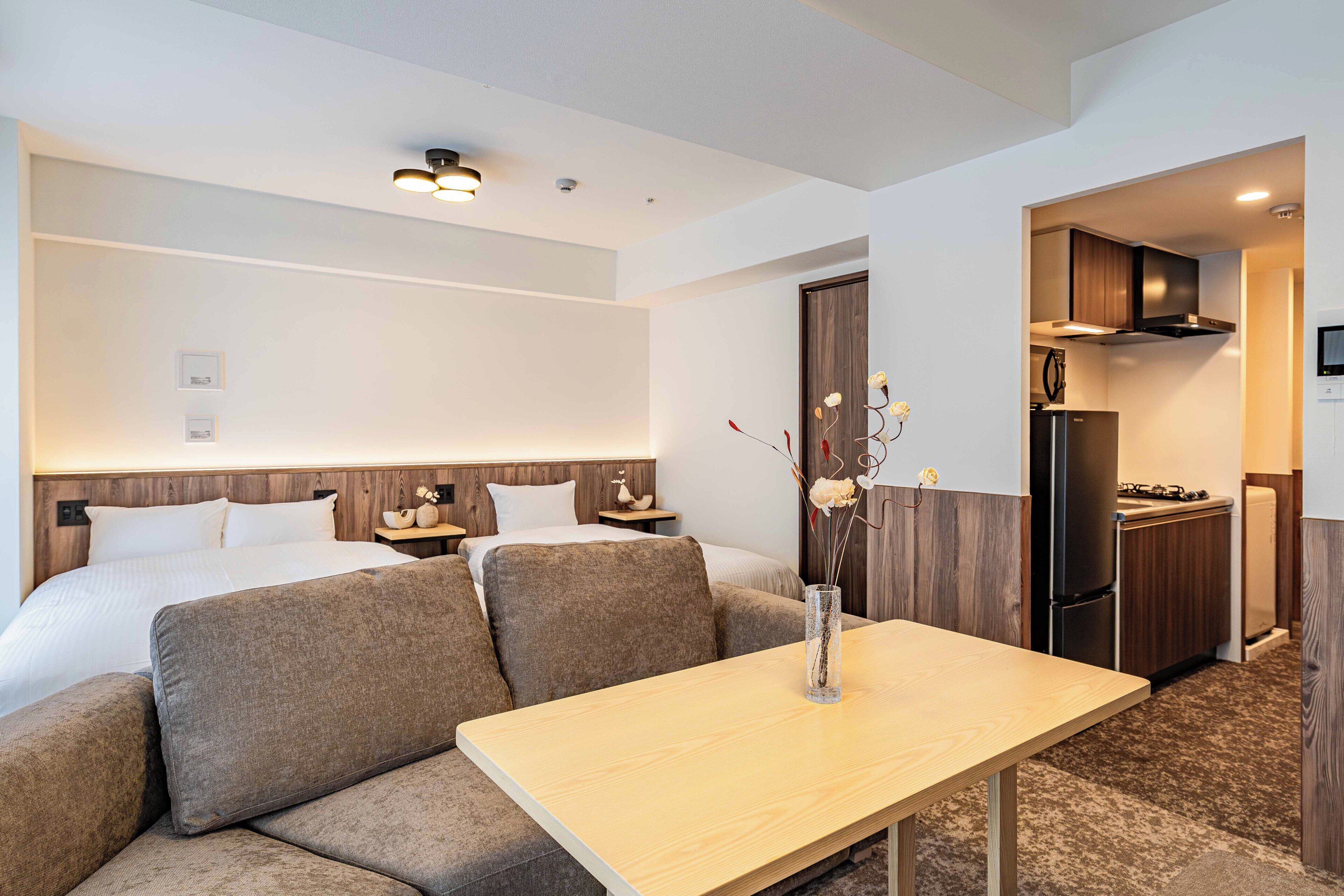 A hotel room with a bed, two sofas, a dining table, a vase with flowers, a kitchen area, and a TV.