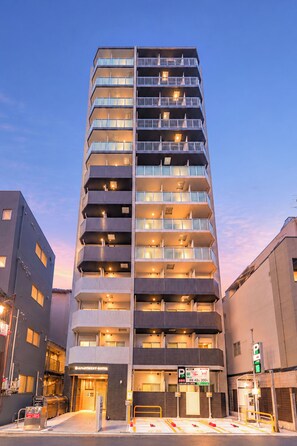 Un hotel de varios pisos con un diseño moderno, que cuenta con balcones en cada planta, una entrada a nivel de calle y un letrero de estacionamiento.