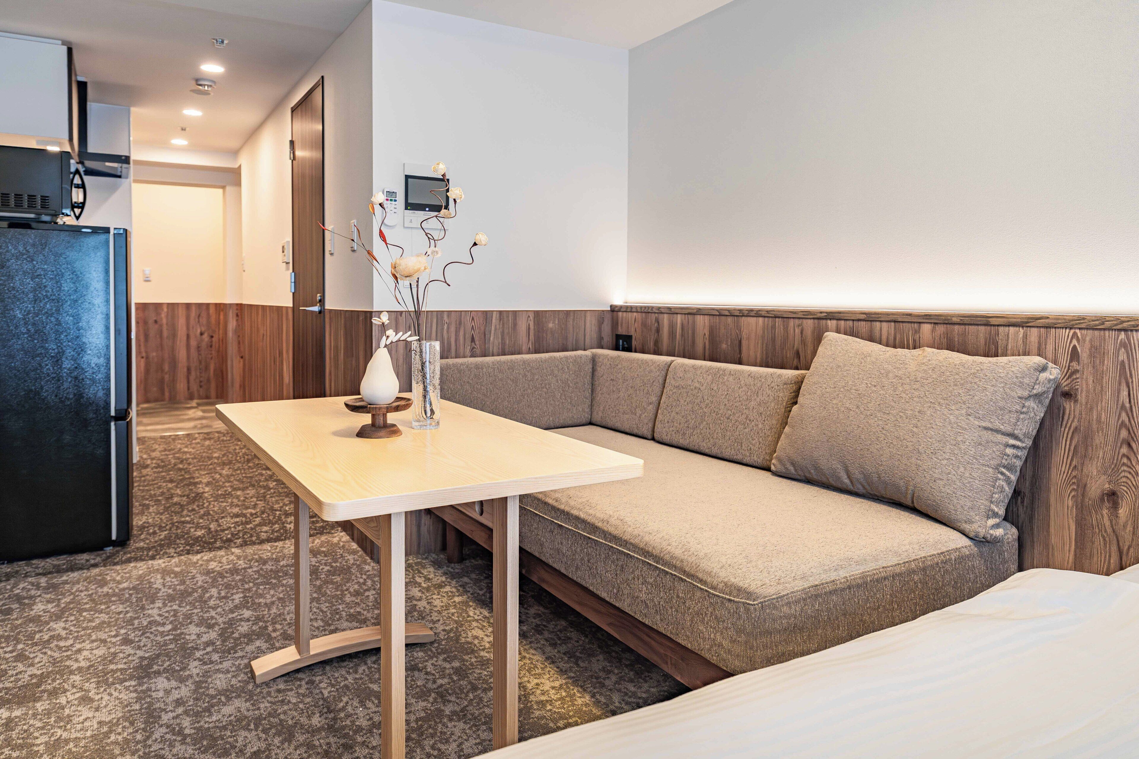 A modern hotel room with a sofa, a small table, a refrigerator, and a wall-mounted control panel.