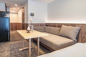 A modern hotel room with a sofa, a small table, a refrigerator, and a wall-mounted control panel.