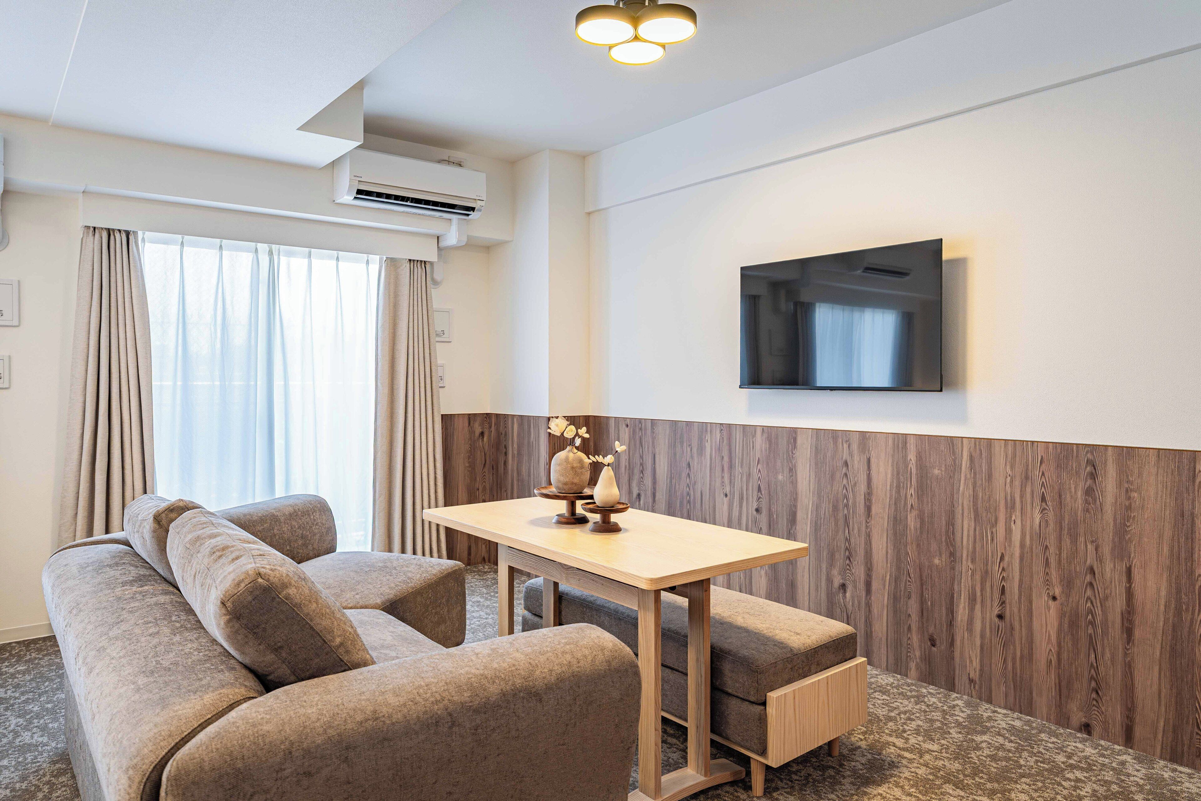 A modern hotel room with a sofa, a small wooden table, a flat-screen TV mounted on the wall, and a window with curtains.