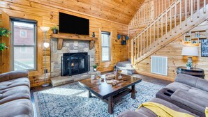 A log cabin interior with a stone fireplace, a flat-screen TV mounted above it, a staircase, and a cozy seating area with a coffee table. - Hot Tub + Game Loft | Located in Douglas Lake Resort | Mountains are Calling (Sevierville)