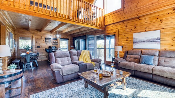A spacious cabin interior with a wooden interior, a balcony, a dining area, and a living room with sofas and a coffee table. - Hot Tub + Game Loft | Located in Douglas Lake Resort | Mountains are Calling (Sevierville)