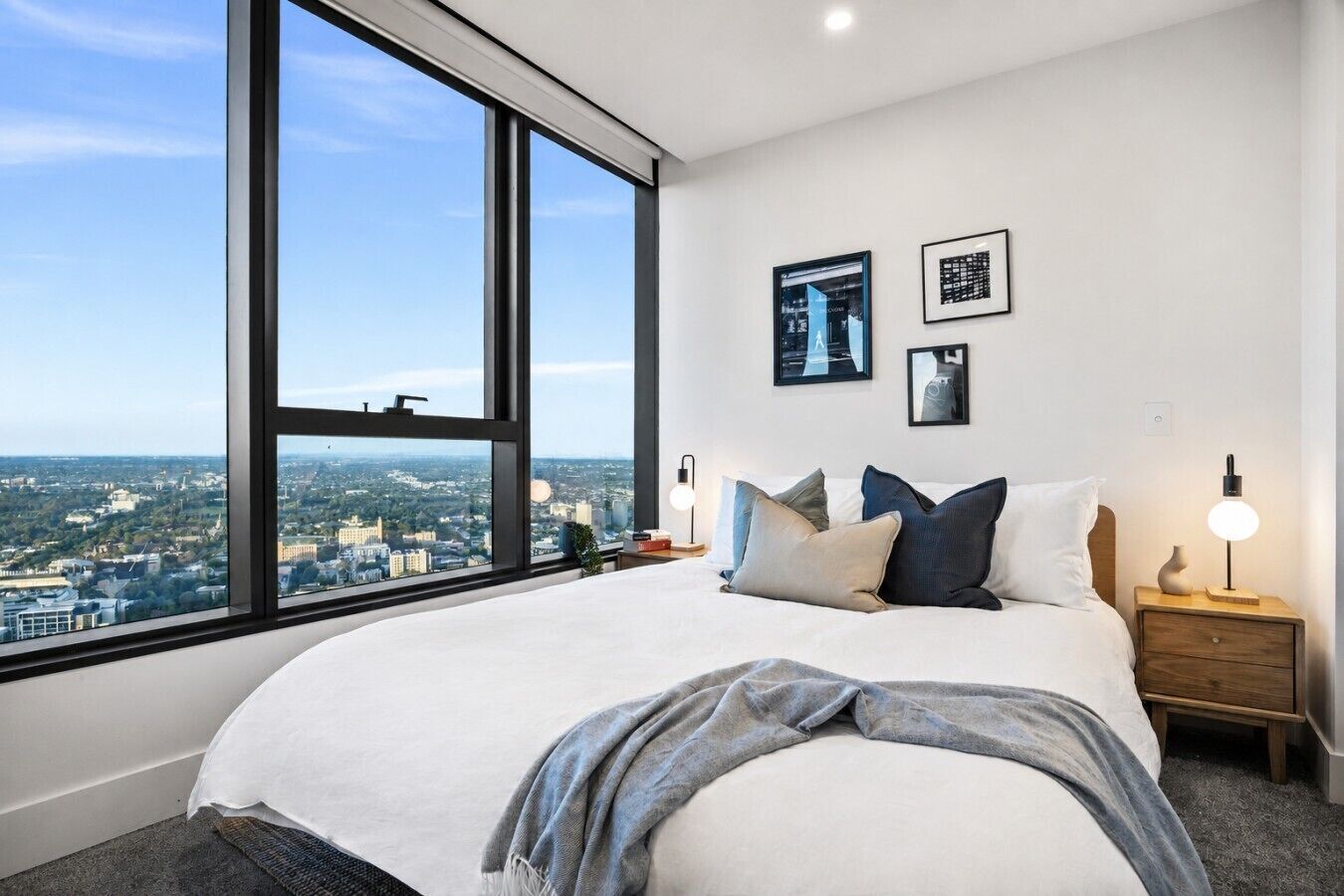 A bedroom with a large window, a bed with pillows, a bedside table with a lamp, and framed pictures on the wall.
