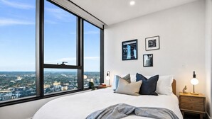 Un dormitorio con un ventanal, una cama con almohadas, una mesita de noche con una lámpara y cuadros en la pared.