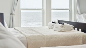 A hotel room with a bed, a view of the sea through large windows, and a neatly made bed with folded towels. - Oceanfront w/ Pool, Panoramic Deck Views, 5BDR | Folly Ocean Breeze by AvantStay (Folly Beach)