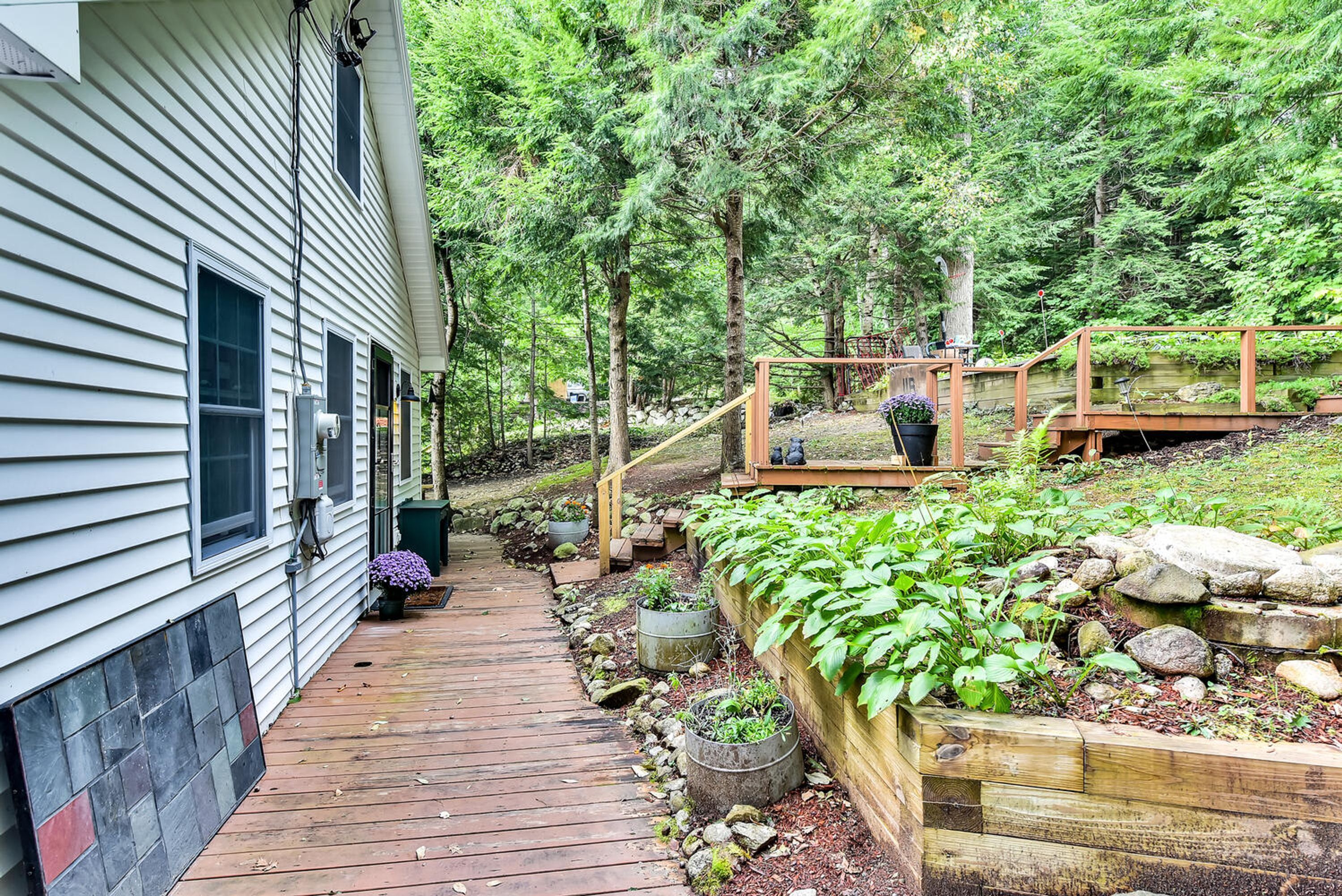 Une terrasse en bois menant à une maison dotée d’un jardin, d’un pont en bois et d’un mur en pierre.