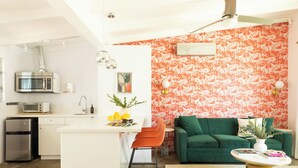A compact kitchen with a microwave, toaster, and refrigerator. A small dining area with a chair and table is adjacent to the kitchen. A green sofa and a coffee table are in the living area, with a patterned wall behind.