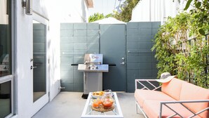 Een terras buiten met een barbecue, een tafel met drankjes en een zithoek met kussens.