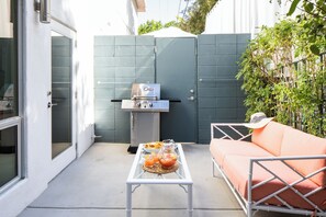 An outdoor patio with a grill, a table with drinks, and a cushioned seating area.
