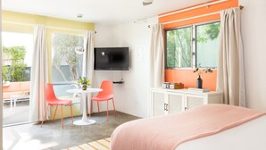 A hotel room with a bed, a TV, a desk with a chair, a window with curtains, and a view of greenery outside.