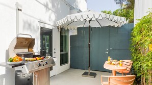 Een terras buiten met een barbecue, een tafel met stoelen en een parasol.