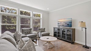 Een moderne woonkamer met een groot raam, een grijze zithoek, een ronde salontafel, een kast met glazen deuren en een schilderij aan de muur.