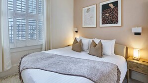 Un lit bien fait, avec du linge de lit blanc et un plaid gris, deux œuvres d’art encadrées au mur et une table de chevet avec une lampe.