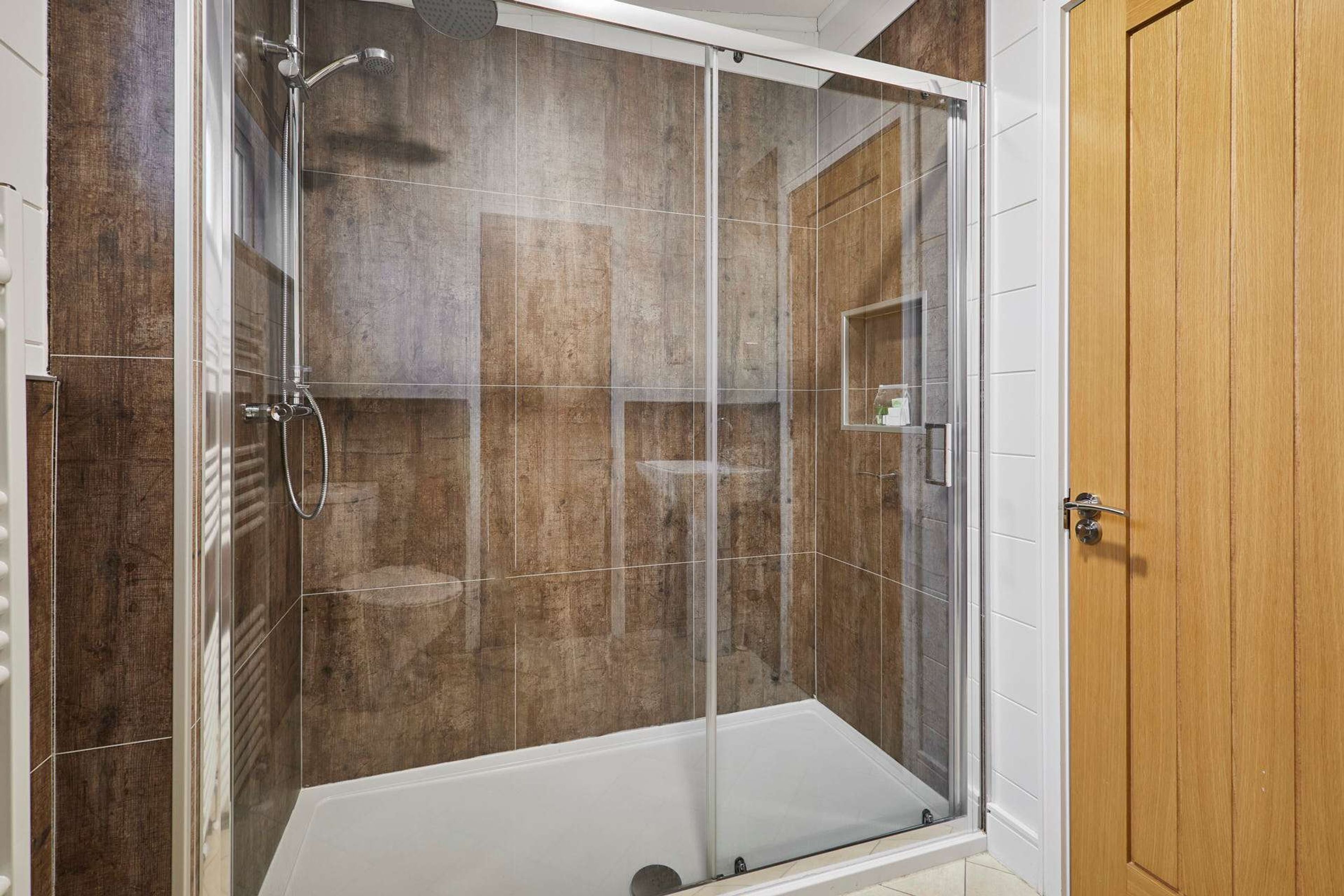 A glass-enclosed shower stall with a white floor, a wooden door, and a tiled wall.