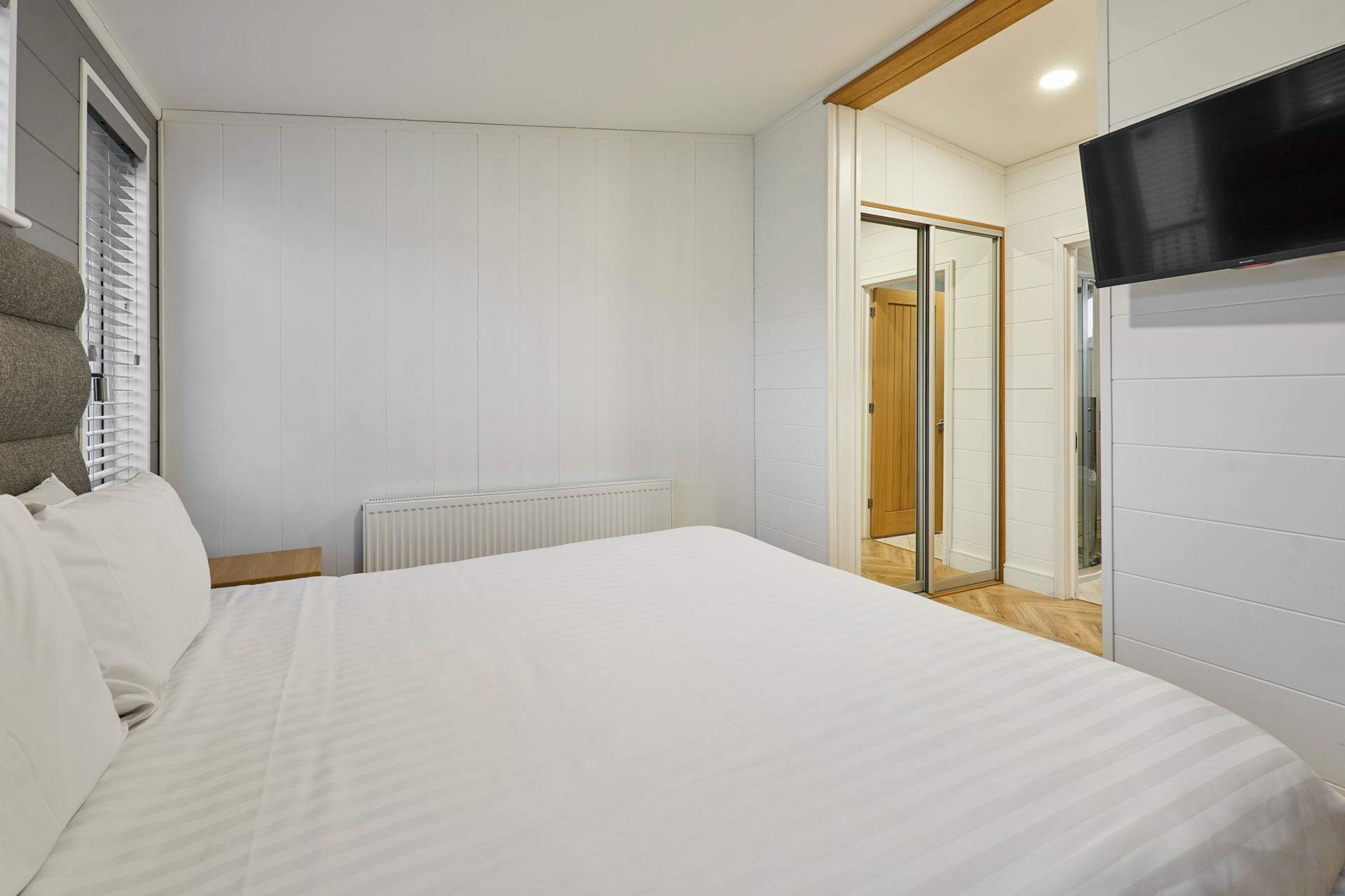 A modern hotel room with a large bed, a flat-screen TV, a radiator, and a sliding glass door leading to a bathroom.