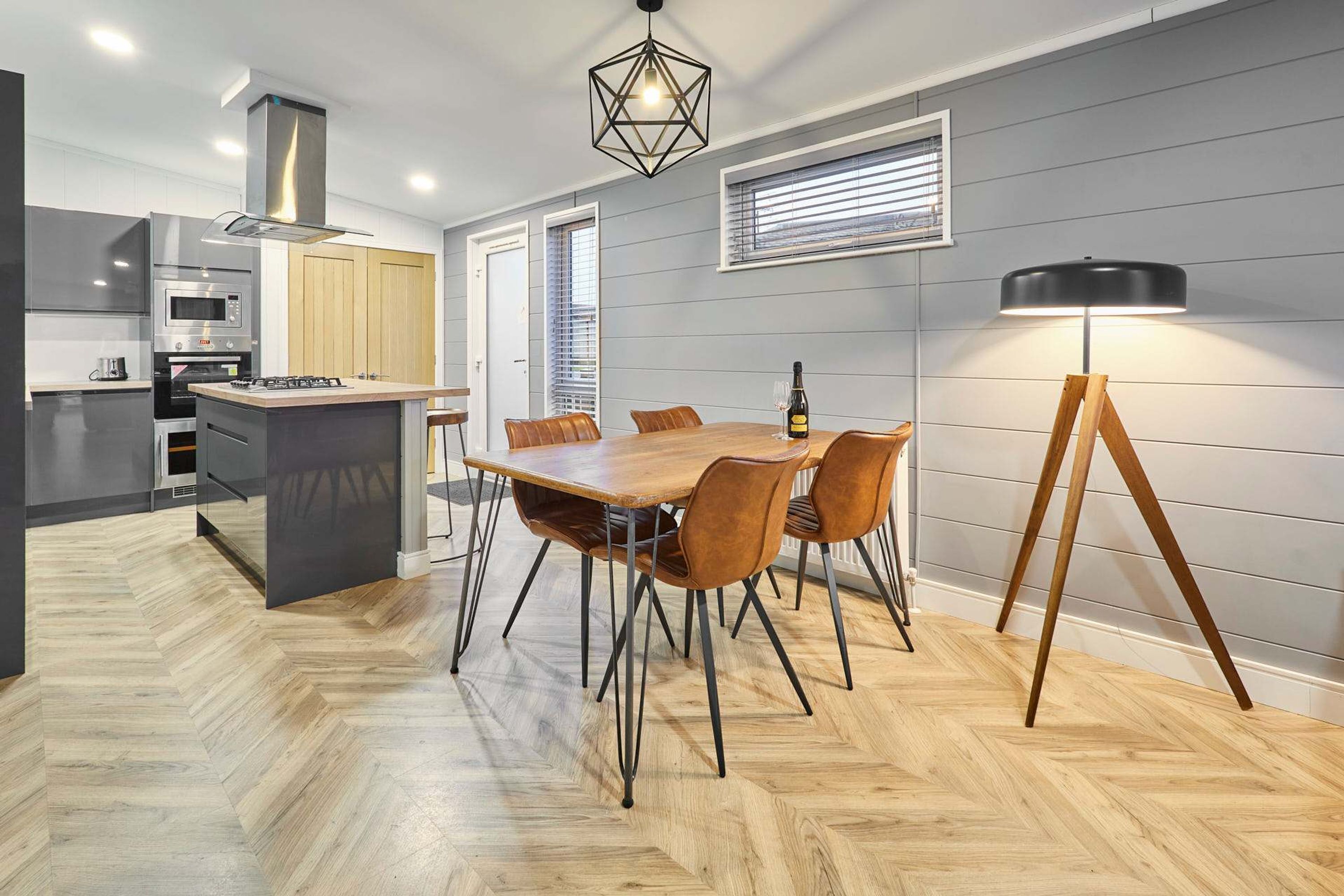 A modern kitchen with a central island, wooden dining table, and chairs, a built-in oven, and a pendant light.
