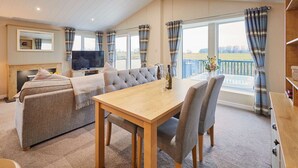 A spacious living room with a sofa, a dining table, and a TV. There is a balcony with curtains and a view of the countryside. - Host Stay Portland 1 (Hexham)