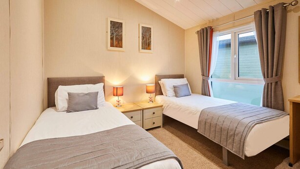 A bedroom with two beds, each with a bedside table and lamp, a window with curtains, and two wall-mounted pictures. - Host Stay Portland 1 (Hexham)