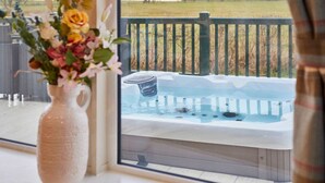 En vy över ett bubbelbad på ett trädäck med ett räcke, en vas med blommor på en fönsterbräda och en skymt av en gräsklädd åker i bakgrunden.