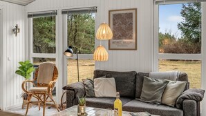 Ett mysigt vardagsrum med en grå soffa, en fåtölj i rotting och ett soffbord med ett ljus och en flaska. Stora fönster ger utsikt över omgivningen.