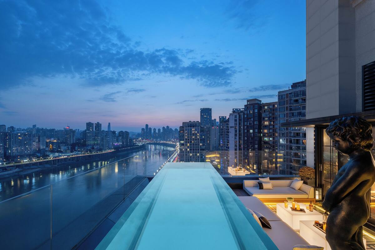 Een dakterras met glazen balustrade, een bank, een beeld en uitzicht op een stadshemelbeeld en rivier bij zonsondergang.