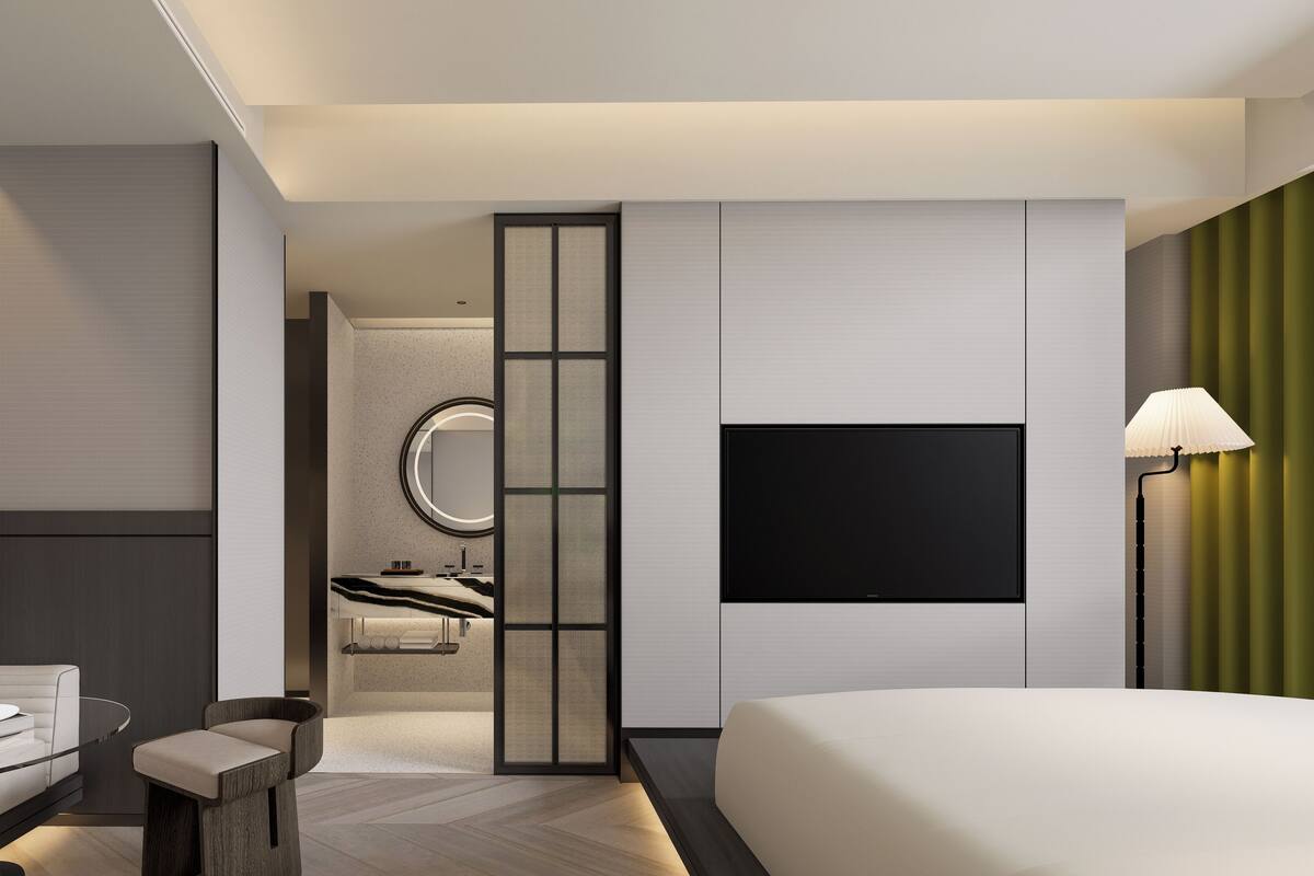 Een moderne hotelkamer met een groot bed, een aan de muur bevestigde televisie, een nachtkastje en een stoel. Een badkamer met een spiegel en wastafel is zichtbaar door een schuifdeur.