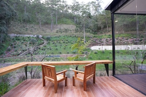 A wooden deck with two chairs and a table overlooking a landscaped garden with a variety of plants and a small pond. - Kiwuki Dalat Resort (Lang Biang - Da Lat)