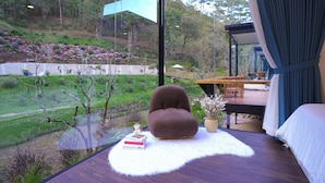 Un dormitorio moderno con un ventanal grande, un sillón marrón mullido, una alfombra blanca y vistas a un jardín y una zona de estar al aire libre.