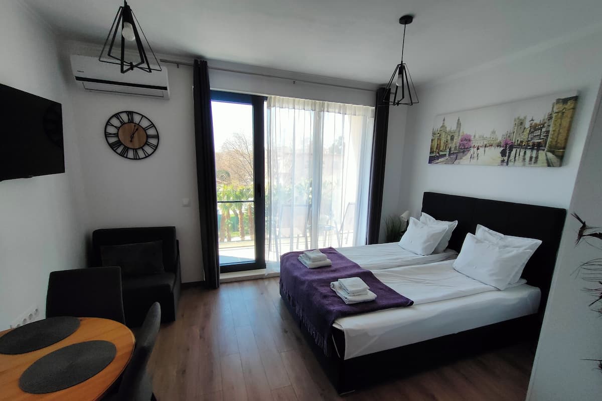 A hotel room with a bed, a desk, a chair, a clock, and a balcony with a view.