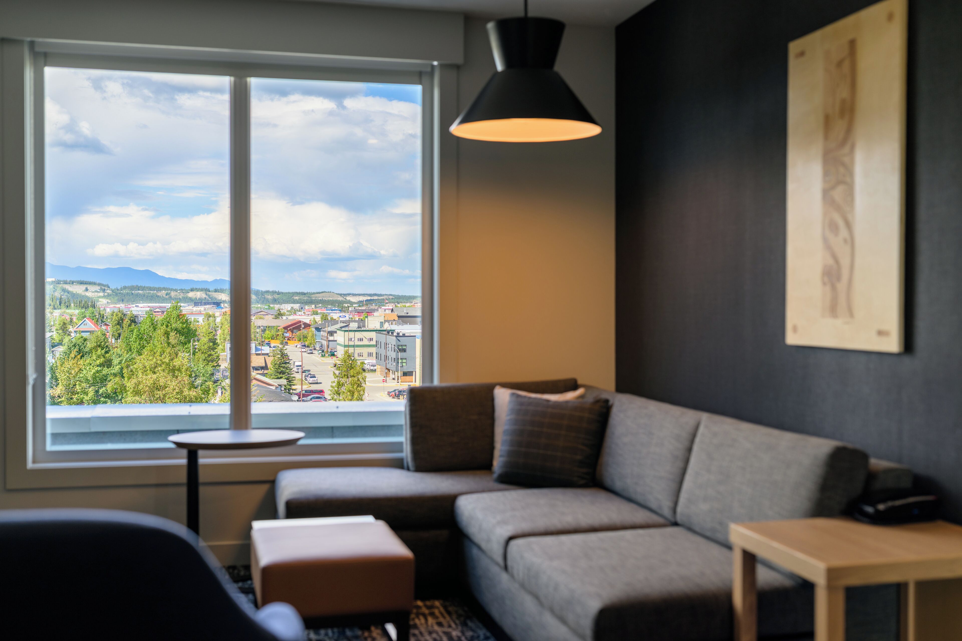 Een moderne woonkamer met een grijze bank, een klein rond tafeltje en uitzicht op een stadsgezicht door een groot raam.