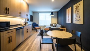 A modern kitchen with wooden cabinets, a built-in microwave, and a dishwasher. There is a dining area with a round table and chairs, and a sofa in the background. - Hyatt Place Whitehorse (WHITEHORSE)