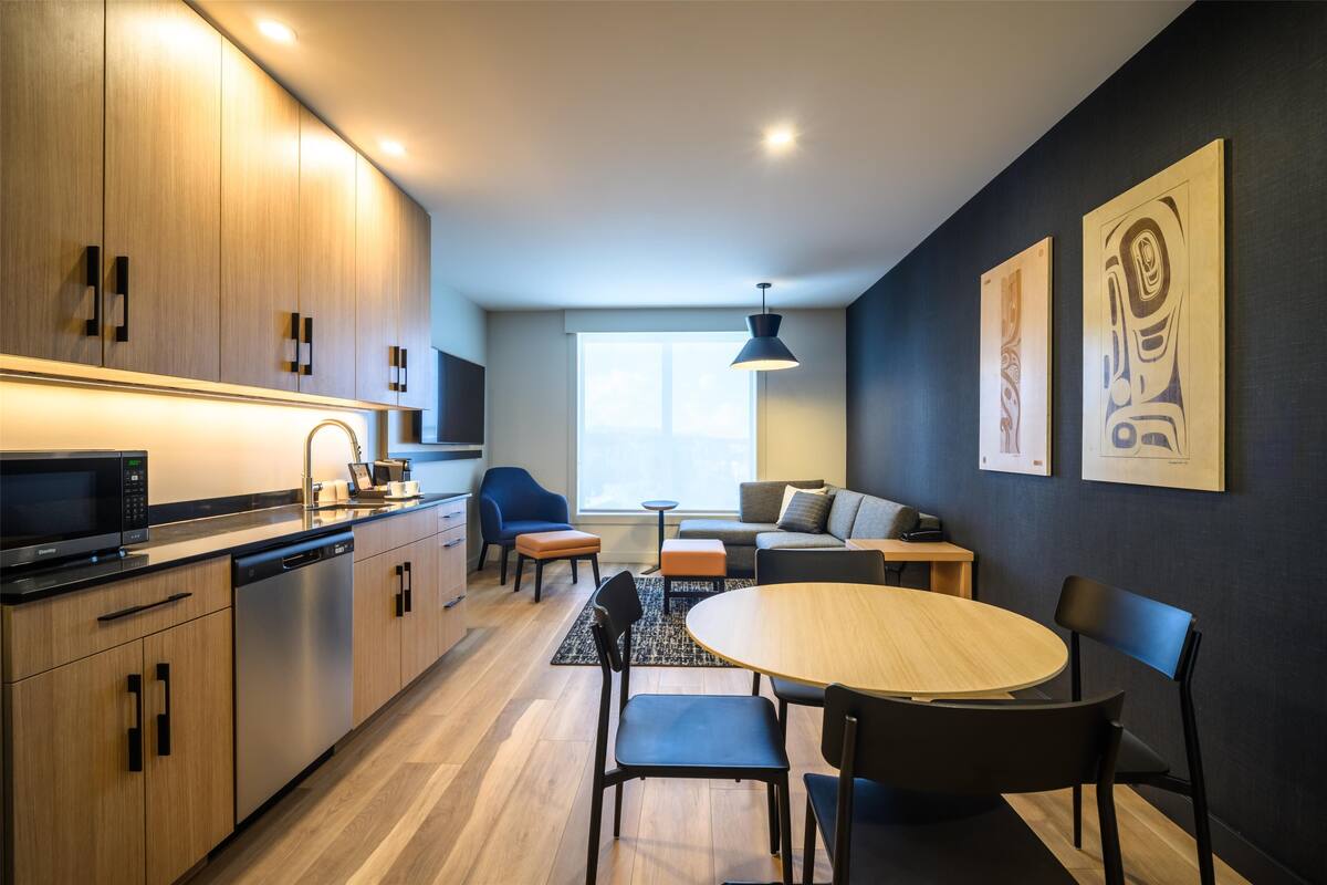 Una cucina moderna con mobili in legno, forno a microonde integrato e lavastoviglie. C'è un'area pranzo con un tavolo rotondo e sedie, e un divano sullo sfondo.
