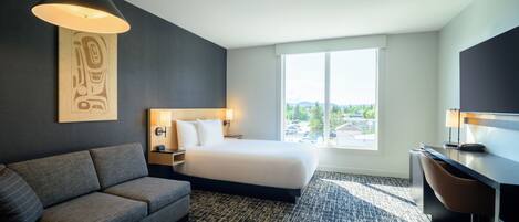 A modern hotel room with a bed, sofa, desk, and large window offering a city view.
