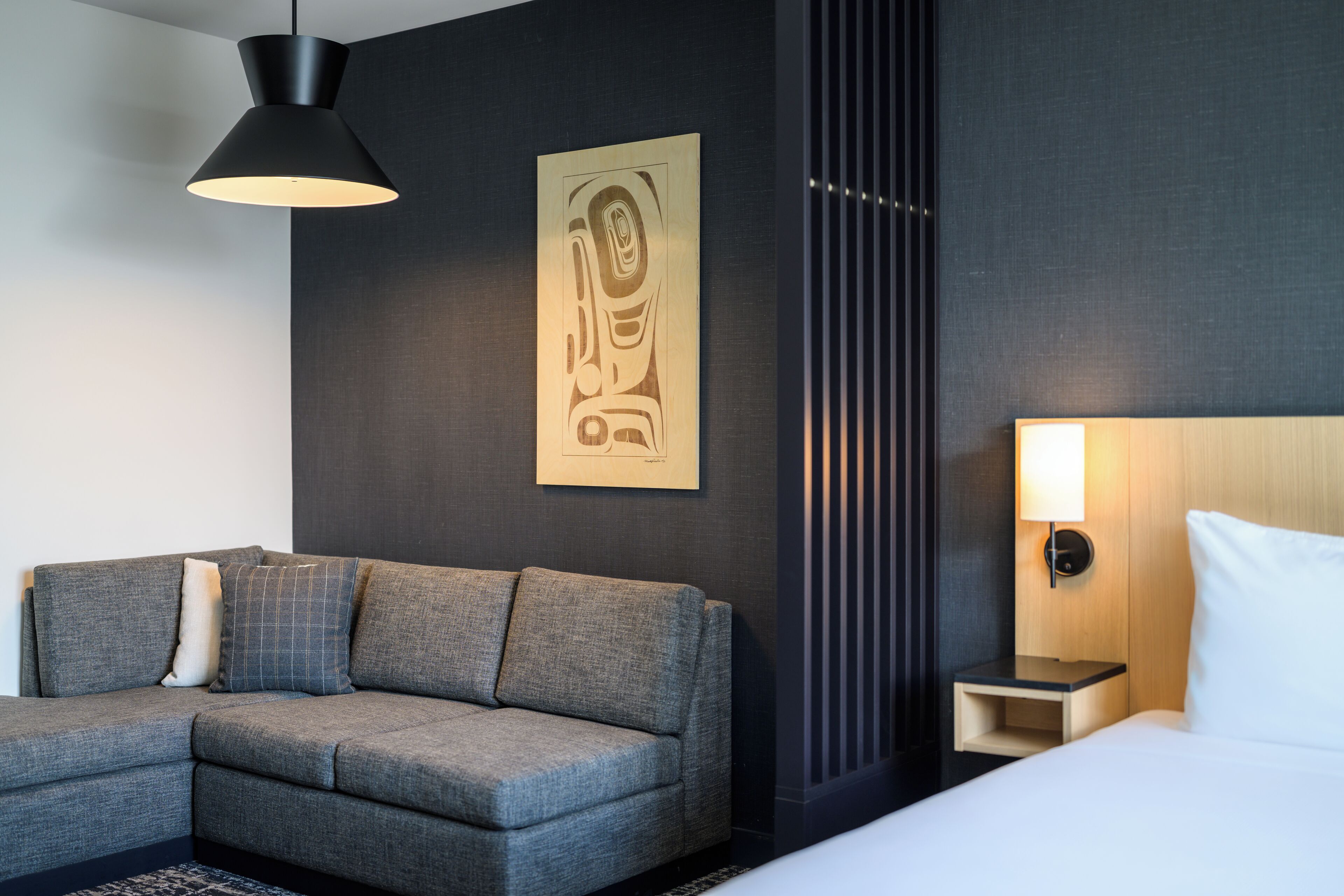 A modern hotel room with a grey sofa, a bed with white bedding, a nightstand, and a wall-mounted lamp.