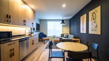 A modern kitchen with wooden cabinets, a built-in microwave, and a dishwasher. There is a dining area with a round table and chairs, and a sofa in the background.
