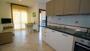 A compact kitchen with white cabinets, a black countertop, and a stove. There is a dining area with a table and chairs, and a TV in the living room. - Three-room apartment Della Rosa Seaside Abruzzo (Alba Adriatica)