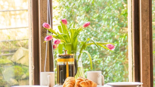 Une table en bois sur laquelle se trouvent un vase de tulipes roses, une cafetière, une tasse et des croissants.