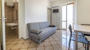 A compact living space with a grey sofa, a small sink, a TV mounted on the wall, and a tiled floor. - Sea View Apartment, Ac Gallipoli (Gallipoli)