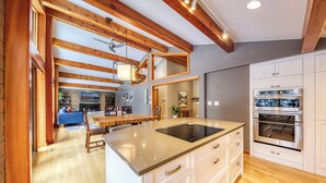 Una cocina moderna con vigas de madera, una isla grande con estufa integrada, armarios blancos y una zona de comedor con sofá azul y mesa de madera.