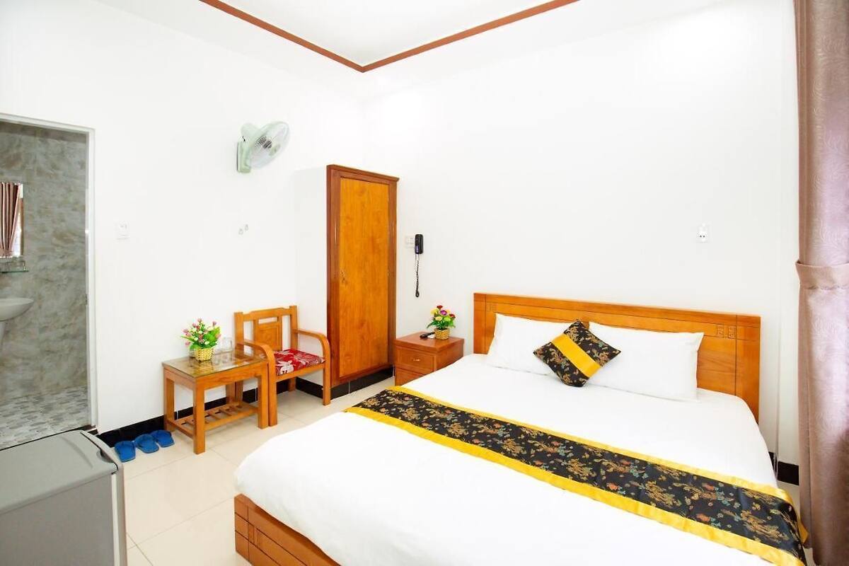 Une chambre d’hôtel comprenant un lit, une table de chevet en bois, une chaise, une petite table, une armoire et un ventilateur fixé au mur.