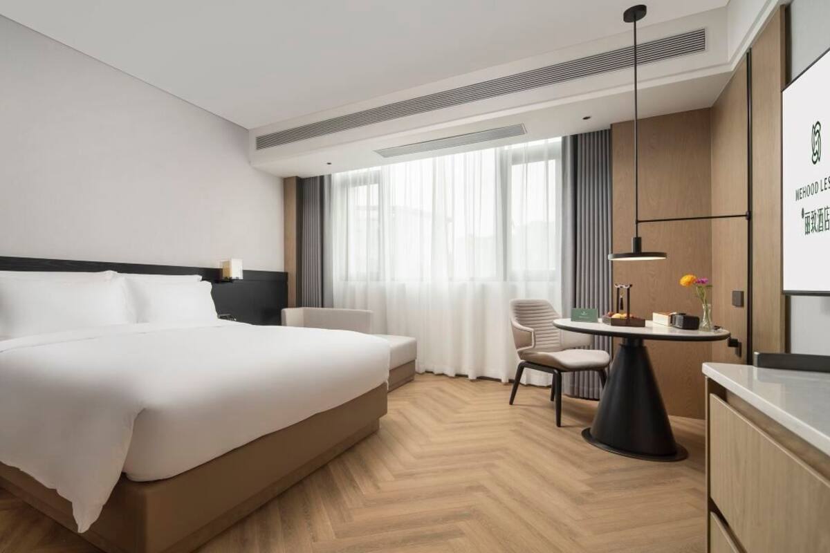 Ein modernes Hotelzimmer mit einem großen Bett, Nachttisch, Lampe, Sessel und einem kleinen Schreibtisch mit Blumen in einer Vase.